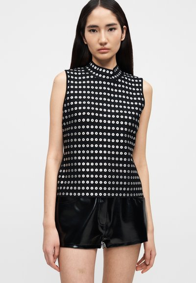 Γυναίκα που φοράει μαύρο τοπ χωρίς μανίκια με λευκά κυκλικά σχέδια και γυαλιστερό μαύρο σορτς, στέκεται μπροστά σε ένα απλό φωτεινό φόντο.