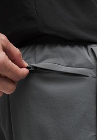 Une main ouvre une fermeture éclair sur un short de sport gris, révélant une poche. La taille est élastique et le t-shirt est noir.