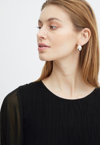 Zilveren druppeloorbellen bij een model. Zwarte geplooide top met een ronde halslijn en transparante lange mouwen. Glad oppervlakdetail op de oorbellen.