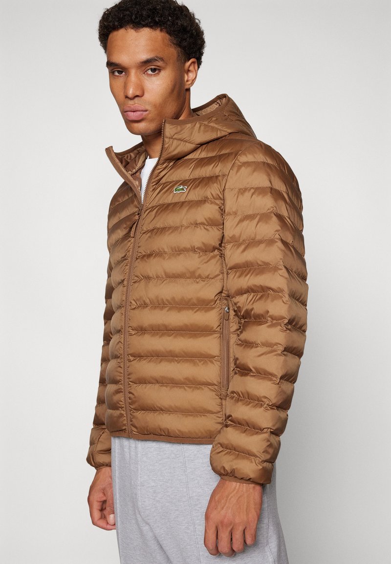 Giacca a vento marrone con cappuccio, caratterizzata da una texture trapuntata orizzontale e un piccolo logo sul petto. Chiusura frontale con zip e tasche laterali.