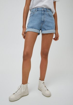 Person iført opsmøgede blå cowboyshorts, hvid t-shirt og hvide høje snørestøvler, stående på en lys baggrund.