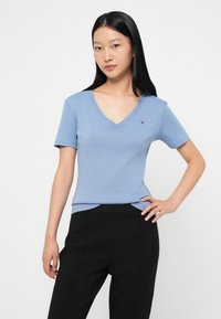 Lyseblå v-hals t-skjorte i et mykt, glatt stoff med korte ermer og en liten logo på venstre bryst. Stylet med svarte bukser.
