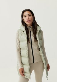 Ljusgrön puffväst med huva, med dragkedja fram och sidofickor. Bärs över en beige åtsittande topp och matchande underdelar.