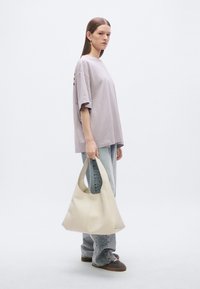 Beige hobo bag made of soft material, featuring a curved top and single strap, paired with a loose-fitting lavender shirt and light blue jeans.