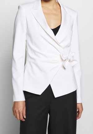 Žena v bílém zavinovacím blejzru zavázaném v pase přes černý top, v černých kalhotách, zobrazená od hrudi po boky.