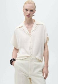 Short-sleeve cream blouse with a collar, button front, and subtle sheen, paired with light cream trousers and a dark bangle on the wrist.