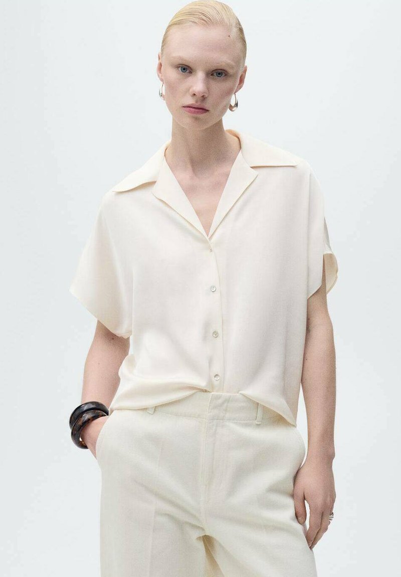 Short-sleeve cream blouse with a collar, button front, and subtle sheen, paired with light cream trousers and a dark bangle on the wrist.