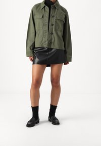 Bavlněná bunda olivově zelené barvy s knoflíky a dvěma náprsními kapsami, doplněná černou koženou mini sukní. Outfit doplňují černé mokasíny a ponožky.