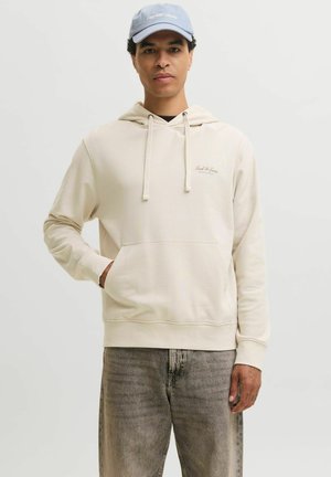 Jeune homme portant un sweat à capuche crème et un jean gris, une main dans la poche et une casquette bleu clair, debout devant un fond clair uni.