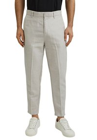 Homme portant un pantalon beige clair sur mesure, une chemise noire et des baskets blanches, debout avec les mains détendues le long du corps.