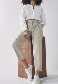 Witte blouse met kanten kraag, beige geblokte broek met riem en witte sneakers. Model staat tegen een houten voetstuk.