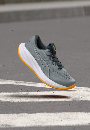 Chaussure de course grise et orange en plein air au-dessus d'un bitume avec des lignes blanches, montrant les détails de la marque et la semelle texturée.