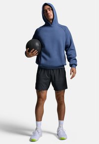 Ein Mann in einem blauen Hoodie und schwarzen Shorts hält einen schwarzen Medizinball. Er steht selbstbewusst vor einem schlichten weißen Hintergrund.