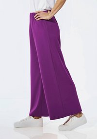 Pantalons larges violets en matériau doux avec une taille élastique, associés à des baskets blanches. Les pantalons présentent une finition lisse et sans texture.