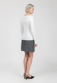 Maglietta bianca a maniche lunghe con scollatura rotonda abbinata a una gonna grigia con pois. Modella che indossa scarpe col tacco nere, vista da dietro.