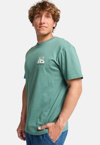 Groen katoenen t-shirt met korte mouwen, ronde hals en een logo aan de voorkant met een abstract ontwerp in beige. Model staat zijwaarts.