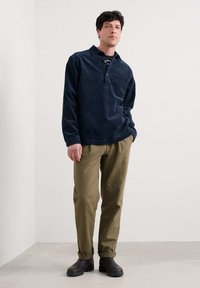 Mornarsko modra kordna srajca z dolgimi rokavi, gumbastim zapenjanjem in žepom na prsih, kombinirana s svetlo olivnimi ožjimi hlačami in rjavimi čevlji brez vezalk.