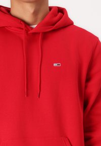 Felpa rossa realizzata in un tessuto morbido, con tasca frontale, coulisse e un piccolo logo ricamato con strisce blu e bianche.