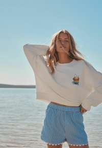 Sweatshirt blanc court avec un graphisme de limonade, associé à un short bleu rayé avec des bords festonnés. Cadre naturel avec de l'eau.