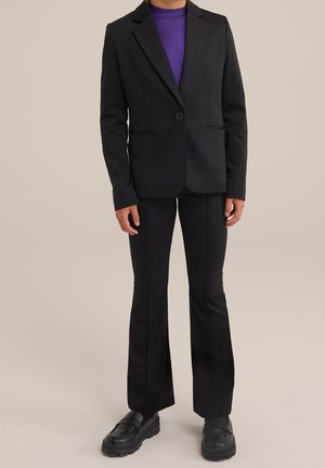 Personne portant un blazer et un pantalon noir sur mesure, un haut violet, et des mocassins noirs épais sur un fond beige uni.