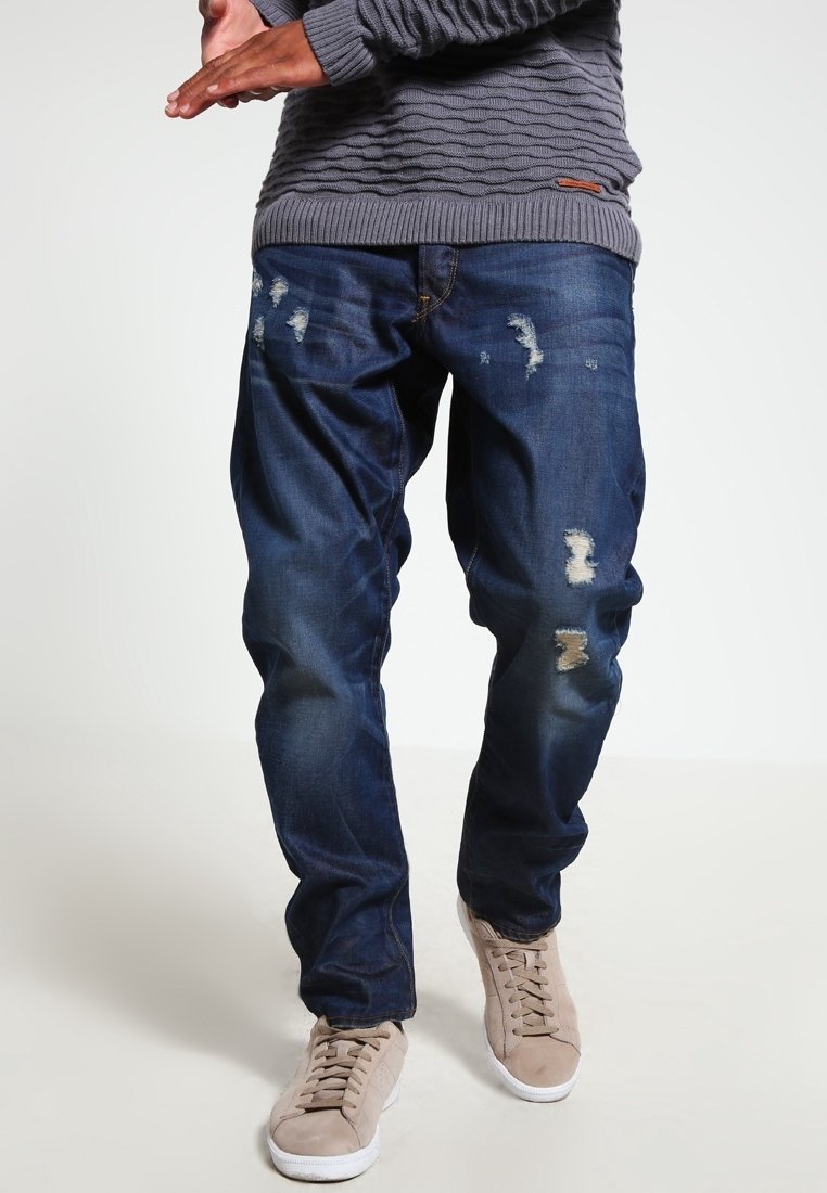 Jeans de mezclilla en un lavado oscuro con parches desgastados y decoloración, combinados con zapatillas de color beige claro y un suéter gris texturizado.