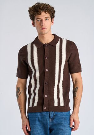 Młody mężczyzna z kręconymi włosami, ubrany w brązowy, krótki sweter z białymi pionowymi paskami, stoi na tle jasnego, jednolitego tła.