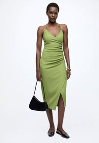 Groene aansluitende midi-jurk met een V-hals en spaghetti-bandjes, voorzien van geplooide details en een split aan de voorkant. Gecombineerd met een zwarte handtas en platte schoenen.