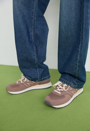 Personne portant un jean bleu et des baskets mauves avec des accents beiges, debout sur un sol vert contre un mur blanc.