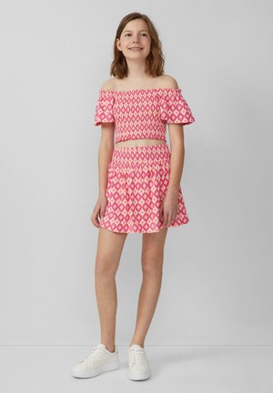 Off-Shoulder, pinkfarbener, gemusterter Crop-Top mit Rüschensäumen, kombiniert mit einem passenden ausgestellten Rock. Getragen mit weißen Sneakers.