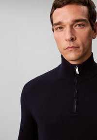 Muž s krátkymi kučeravými vlasmi, oblečený v čiernom svetri na zips, stojaci pred jednofarebným svetlosivým pozadím.