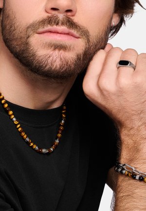 Uomo barbuto che indossa una camicia nera, collana di perle occhio di tigre, braccialetto abbinato, anello d’argento con pietra nera e piccolo orecchino a cerchio.