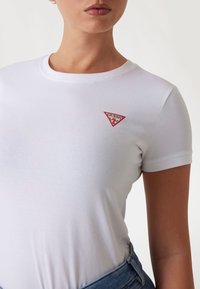 T-shirt bianca in cotone con scollo rotondo, caratterizzata da una toppa rossa a forma di triangolo con il logo 'GUESS' sul lato sinistro del petto. Maniche corte e design aderente.