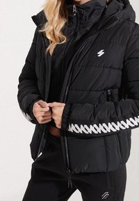 Chaqueta acolchada negra con capucha, cierre frontal con cremallera, patrón de zigzag blanco en las mangas y puños elásticos. El material parece suave y cálido.