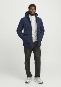 Marinblå pufferjacka med huva, som har en frontdragkedja, två stora fickor och en figursydd design, bärs över en ljusgrå sweatshirt och mörka jeans.