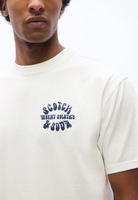 Νεαρός άνδρας φορώντας ένα εκρού μπλουζάκι με μπλε σκούρο κείμενο «Scotch Desert Solstice & Soda» στην αριστερή περιοχή του στήθους.