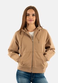  sweat-shirt zippé beige en tissu doux, avec une capuche, des cordons de serrage, des poches kangourou et un logo brodé sur la poitrine.