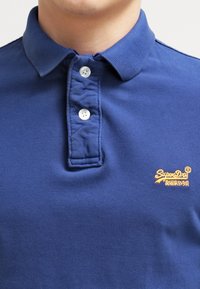 Marinblå polo skjorta i strukturerad tyg med knappad krage, tre knappar och en liten gul logotyp på vänster bröst.