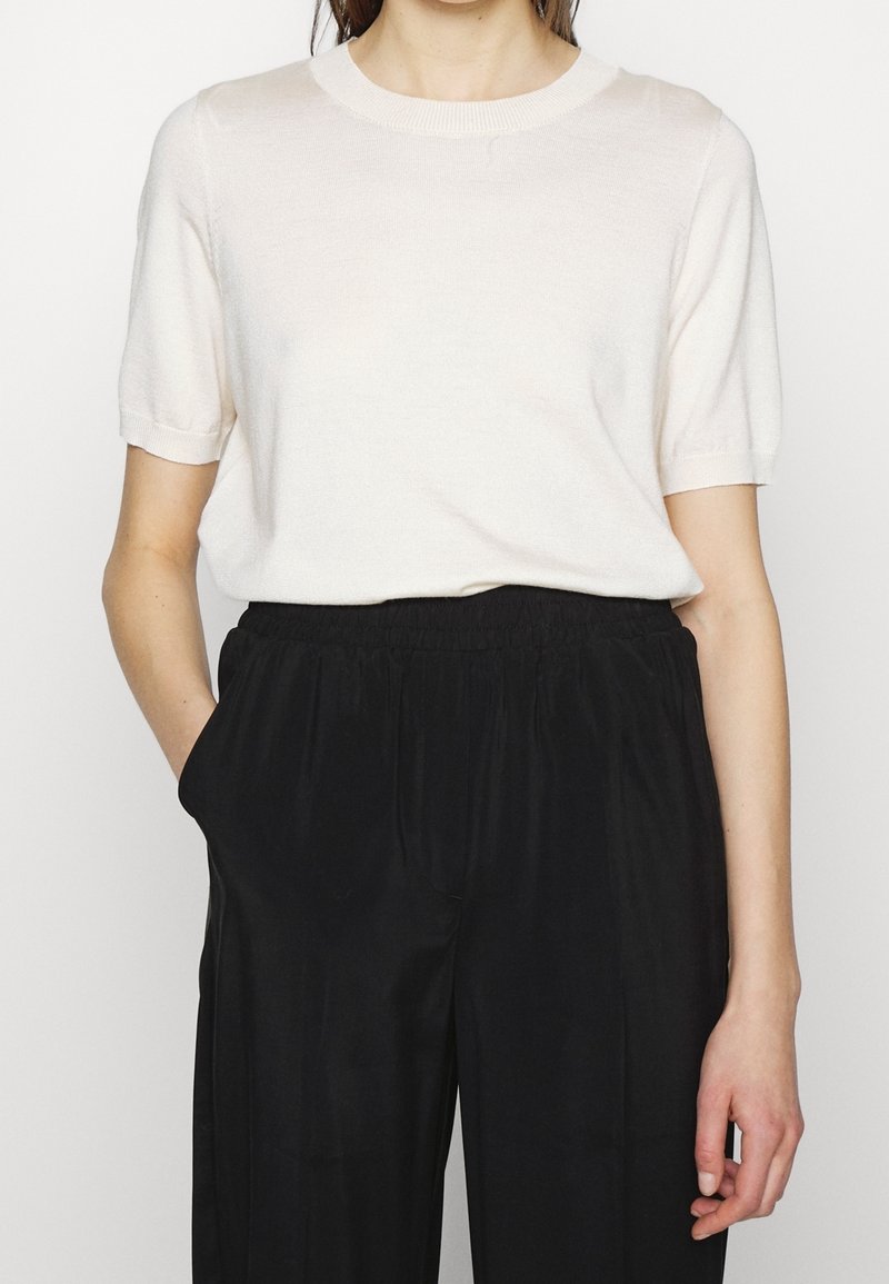 Personne portant un haut blanc à manches courtes rentré dans un pantalon noir ample, debout avec une main dans la poche devant un fond uni.