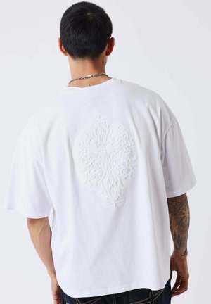 Man draagt een los wit T-shirt met een reliëf met een sierlijk ontwerp op de rug, staand met de rug naar een effen achtergrond.