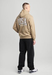 Beige hoodie with "USA" and "NEW YORK CITY" printed in large text on the back. Paired with black pants and white sneakers.