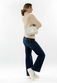 Borsa a tracolla beige trapuntata con hardware rettangolare argento, indossata a bandoliera. Modella in maglione crema e jeans blu, in piedi di profilo.