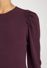 Ribbade trikåtoppar i djup burgundy med rund halsringning och puffärmar, som visar upp textilens struktur och en figurnära design.