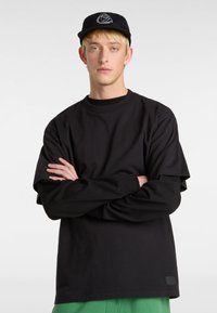Schwarzes langärmliges T-Shirt mit lockerer Passform und Rundhalskragen, unter einem schwarzen Hoodie getragen. Das Modell trägt eine schwarze Mütze mit einem weißen Logo.