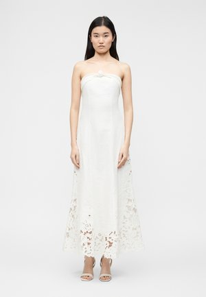 Donna che indossa un abito bianco senza spalline con dettagli in pizzo floreale all'orlo, in piedi dritta contro uno sfondo chiaro e semplice.