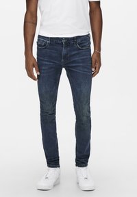 Sötétkék, slim-fit farmernadrág, enyhe kopással �és öt zsebes tervezéssel. Fehér sportcipővel párosítva, lapos elülső résszel és egyenes lábakkal.