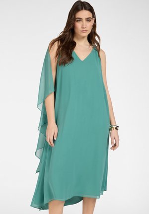 Donna con capelli lunghi castani che indossa un vestito turchese senza maniche, fluente, con schiena a strati in chiffon e braccialetti dorati, in piedi davanti a uno sfondo semplice.