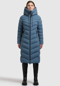 Gara, ar kapuci, piepūsta jaka maigā zilā krāsā ar chevron izšuvumu, divām ar rāvējslēdzējiem aprīkotām kabatām un priekšējo rāvējslēdzēju. Valkāta ar melniem ceļgalu augstiem zābakiem.