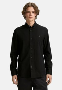Camisa negra de mangas largas con botones, hecha de una tela texturizada, con un cuello, botones frontales y un pequeño logo en el lado izquierdo del pecho.