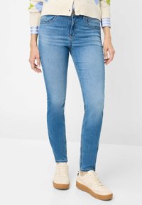 Jean skinny bleu clair en denim, avec poches classiques et légère décoloration. Associé à des baskets beige et un cardigan crème texturé.