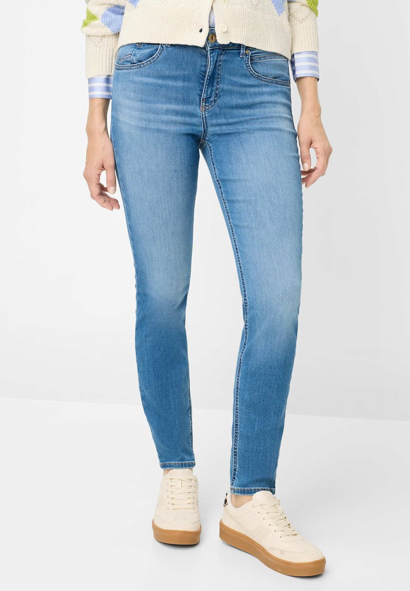 Jean skinny bleu clair en denim, avec poches classiques et légère décoloration. Associé à des baskets beige et un cardigan crème texturé.
